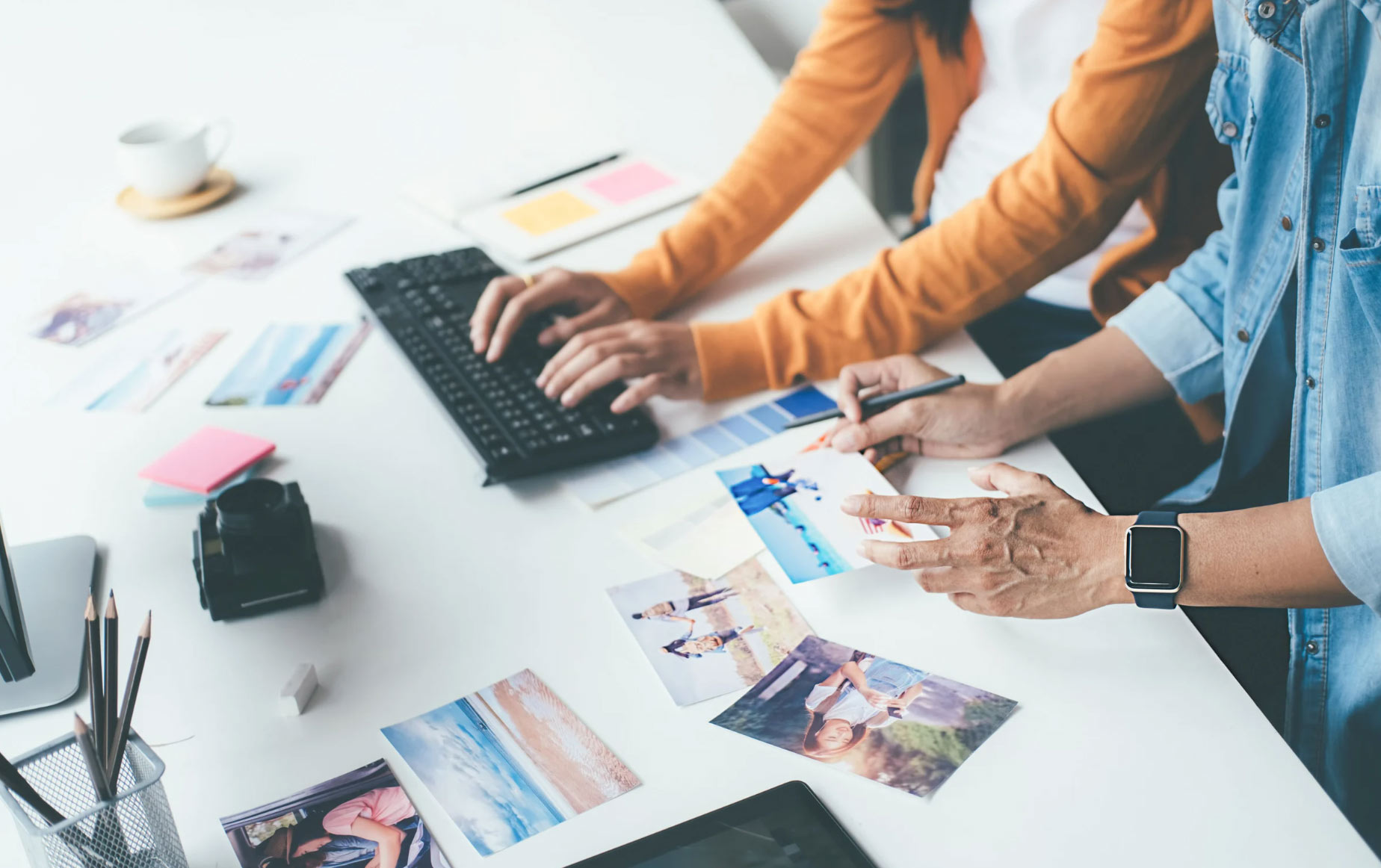 bk-bg-img Two people editing and sorting photographs at a desk with a keyboard, camera, and photos spread out.