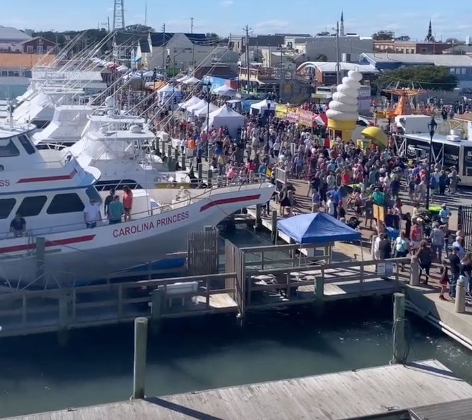 Crowds gather at a marina festival with boats, tents, and a large inflatable ice cream cone visible.