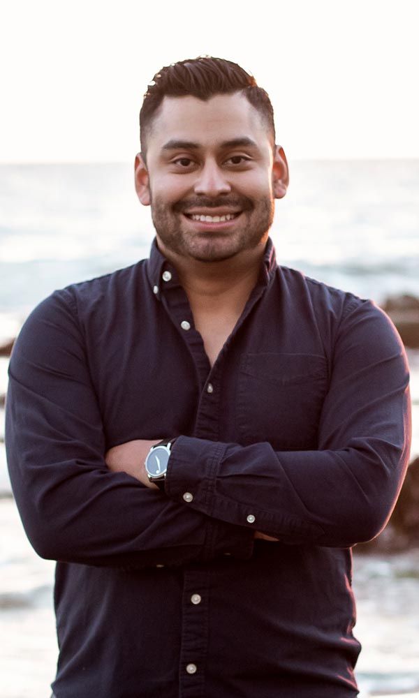 Smiling man with arms crossed, wearing a navy shirt, standing outdoors by the water.