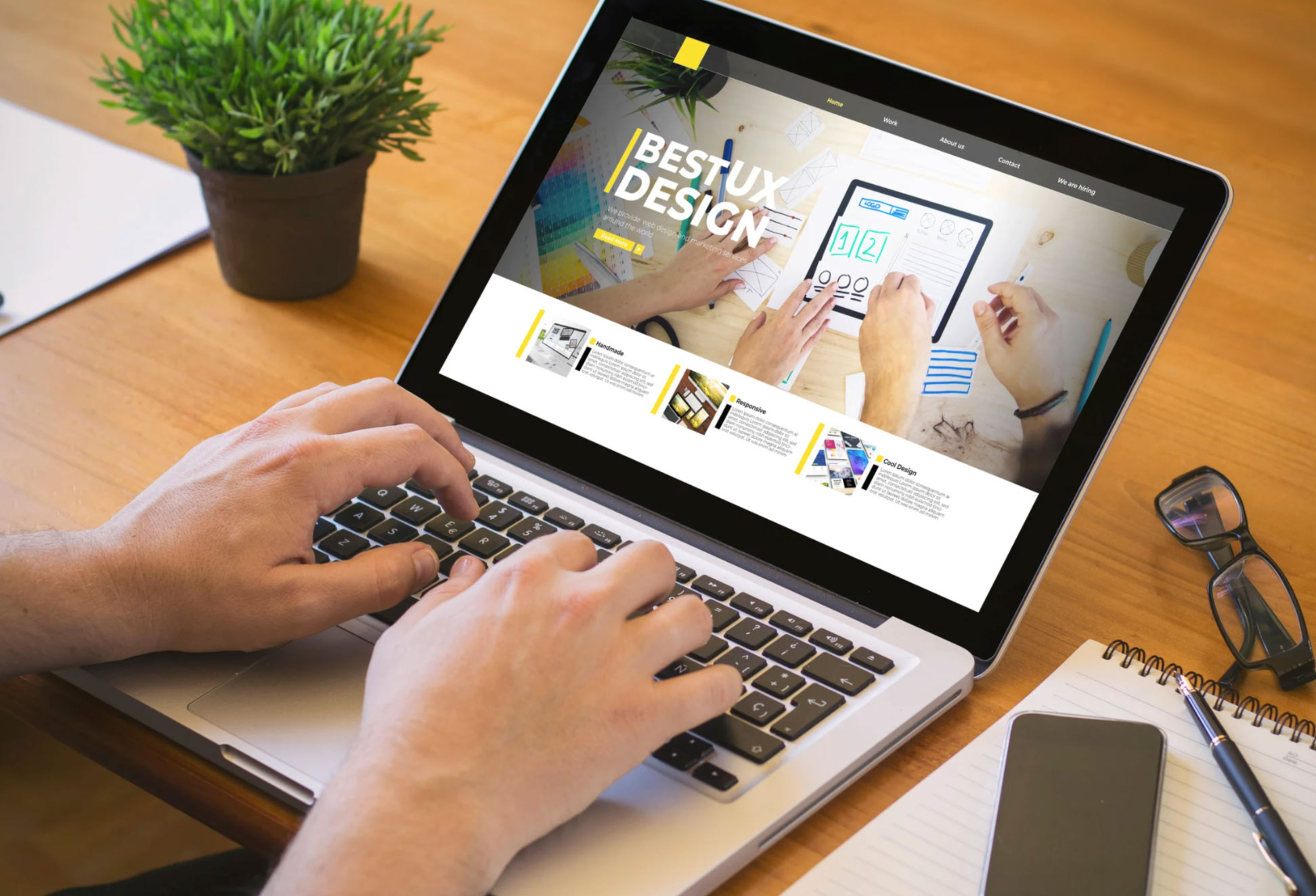 web-des-img Person using a laptop displaying a "Best UX Design" webpage, with a notebook and plant on the desk.