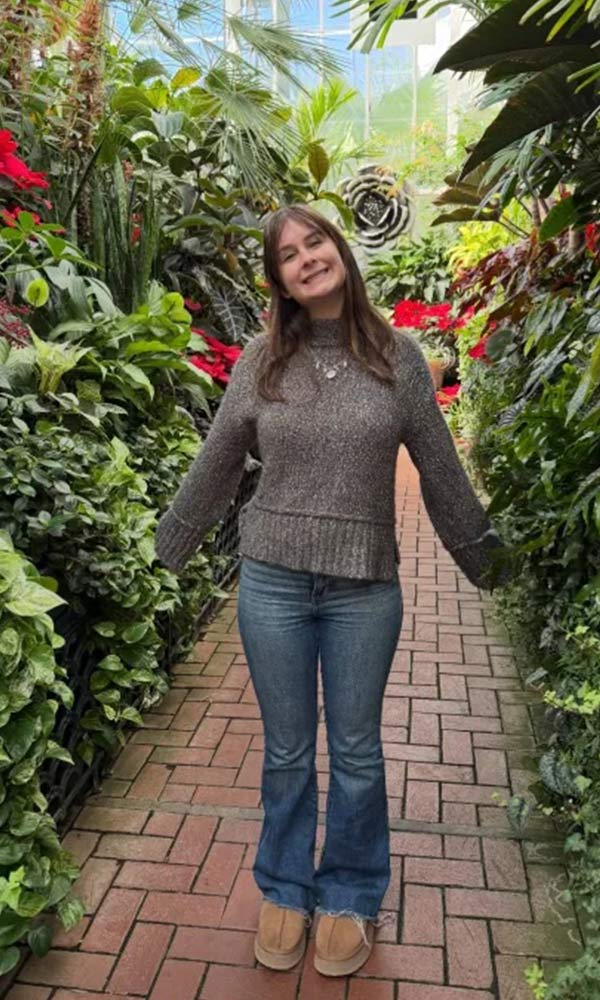 Woman smiling and standing on a brick path surrounded by lush green plants and red flowers in a greenhouse.