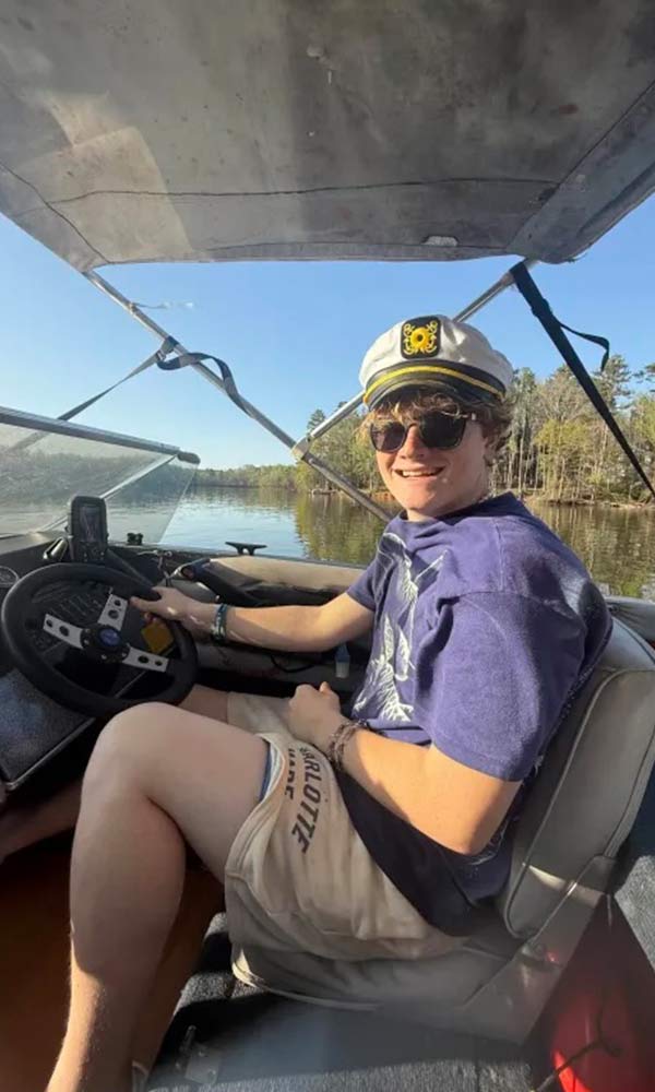 Smiling person in a captain’s hat drives a boat on a sunny day, surrounded by calm water and trees.