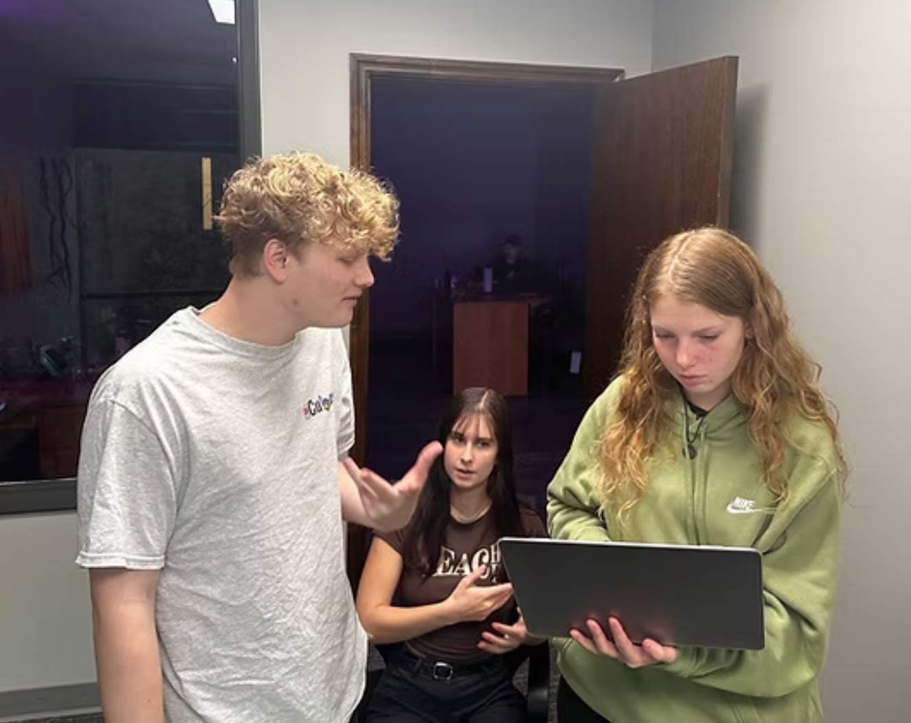 why-ohcool-img Three young people, two standing and one sitting, look at a laptop in a room with gray walls and an open door.